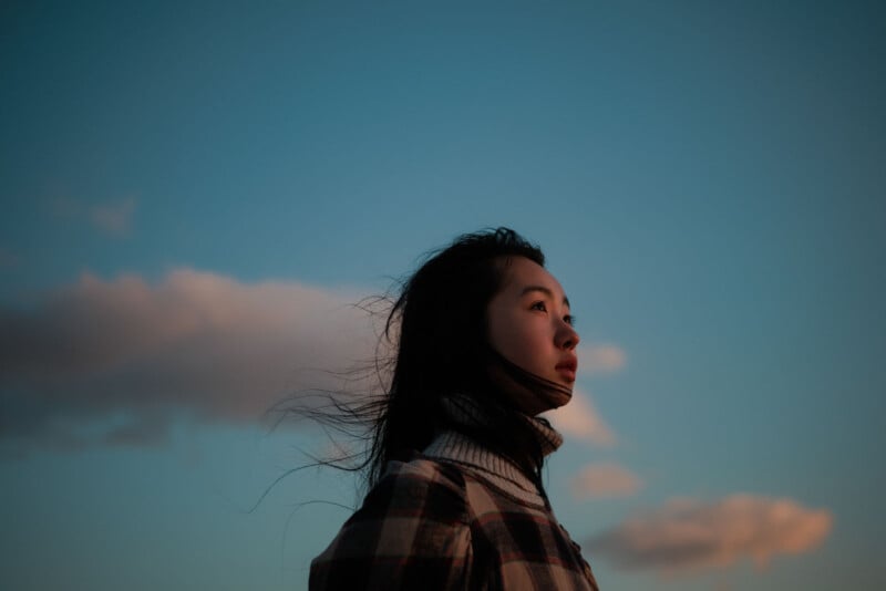 A person with long hair wearing a plaid jacket looks thoughtfully into the distance, with the sky and clouds in the background at sunset.