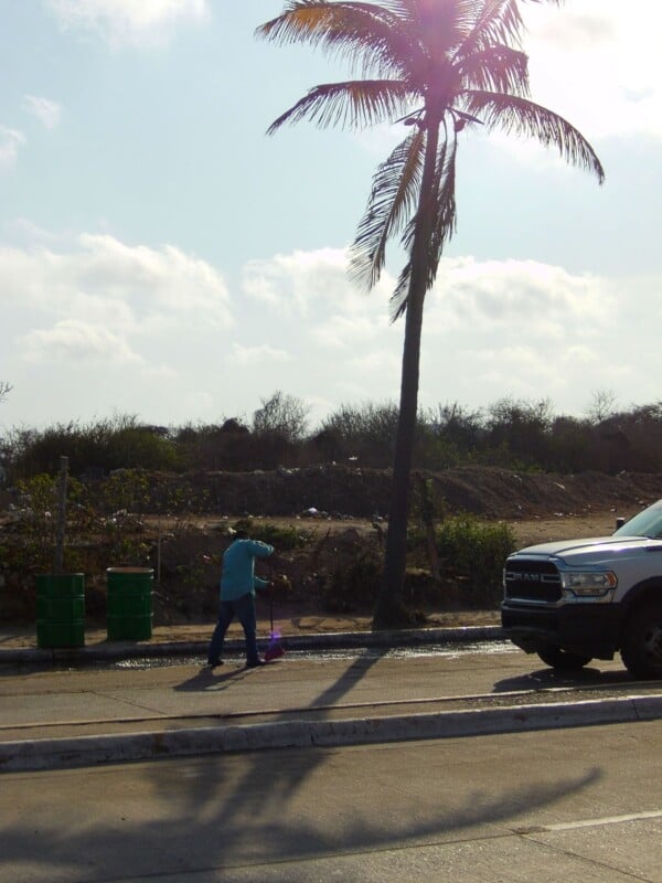 A person sweeps the street near a palm tree and a parked white pickup truck under a partly cloudy sky. There are green garbage bins and shrubbery in the background.