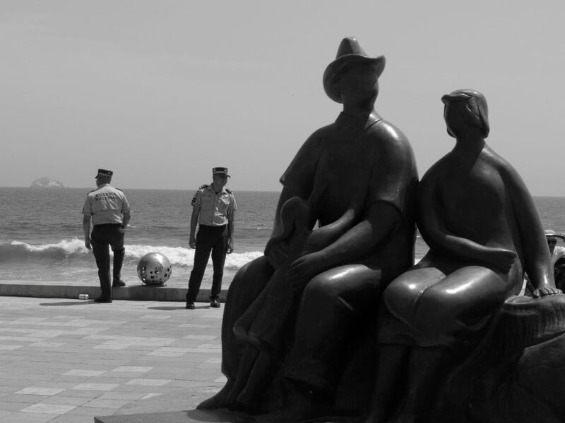 Two police officers stand by the ocean, while a large statue of a seated couple faces the water in the foreground. The scene is in black and white, with waves and an island visible in the background.