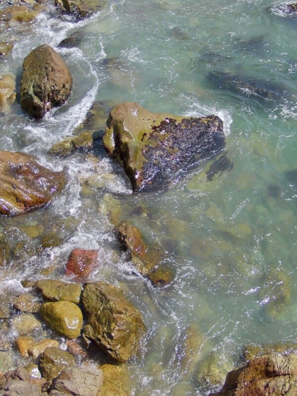 Clear turquoise water flows around mossy rocks and stones along a shoreline, with gentle waves creating white foam at the edges.