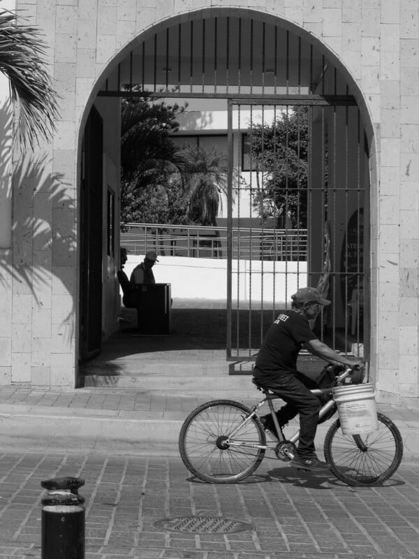 A man rides a bicycle with a bucket on the front along a street, passing by a large arched gateway. Inside the gateway, two people sit in the shade, with trees and buildings in the background. The scene is in black and white.