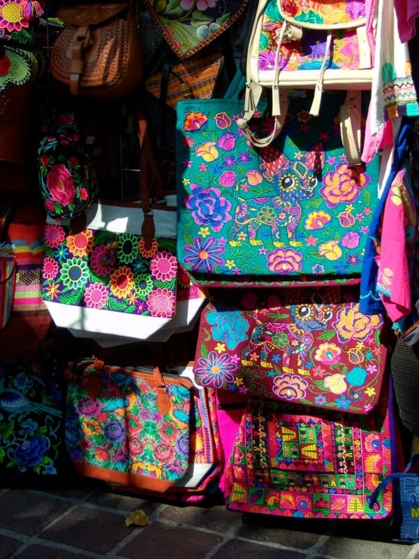 Colorful embroidered bags with vibrant floral and geometric patterns are displayed for sale at an outdoor market stall, hanging against a wall and shining in the sunlight.