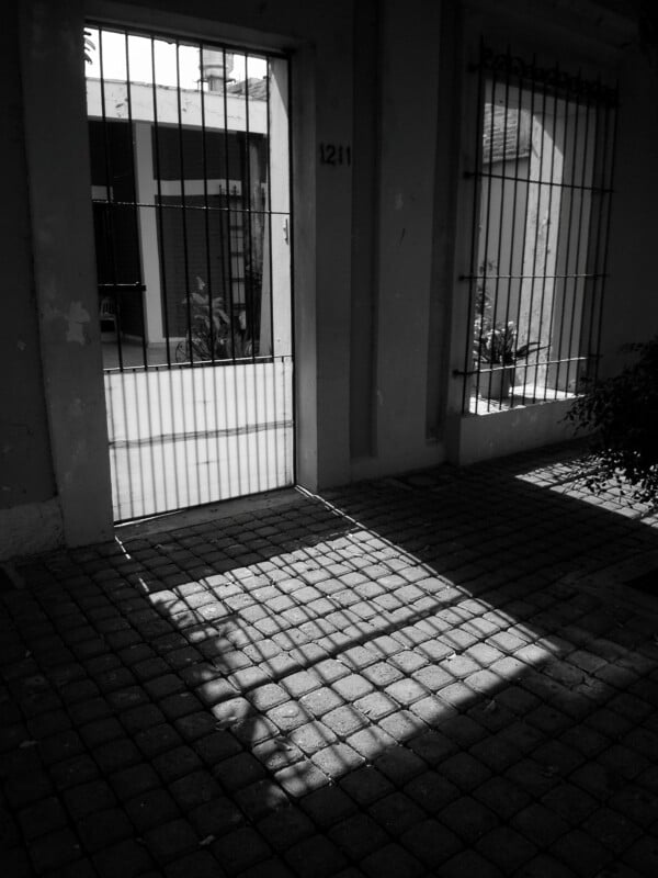 Sunlight streams through an open wrought iron gate, casting distinct shadows on a tiled pathway. The scene is in black and white, highlighting the contrast between light and shadow.