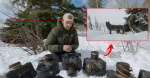 A person in winter clothing kneels in snow behind several cameras. An inset shows a black dog-like animal standing in deep snow, indicated by a red arrow. Snowy trees and landscape fill the background.