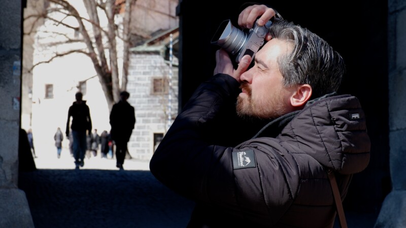 Man in a black jacket takes a photo in a shadowed area with a camera. Sunlit street and blurred figures are in the background.