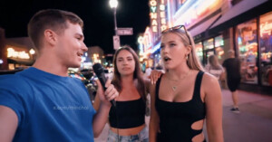 A man in a blue shirt holds a microphone, interviewing two women on a busy street at night with neon lights and storefronts in the background. The woman on the right looks surprised, while the other smiles.
