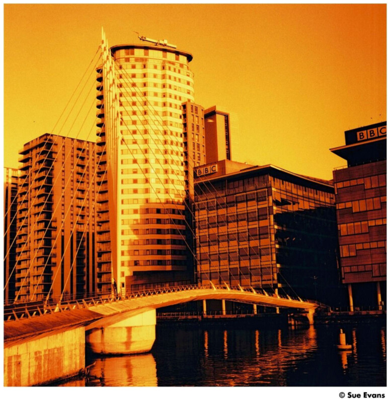 A pedestrian bridge spans a river, leading to modern glass and steel buildings, including two labeled "BBC." The image has a strong orange-yellow tint, creating a warm, dramatic effect. Photo credit: Sue Evans.
