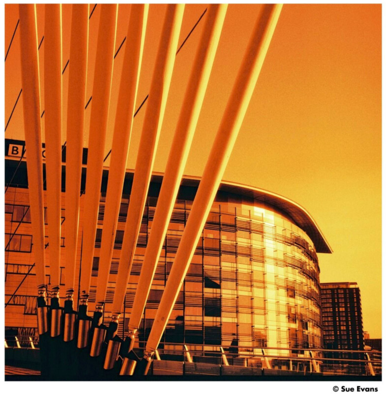 A modern building with a curved glass facade is seen behind large, angled yellow support cables. The photo has a strong orange-yellow filter, giving it a warm glow. The BBC logo is visible on the building.