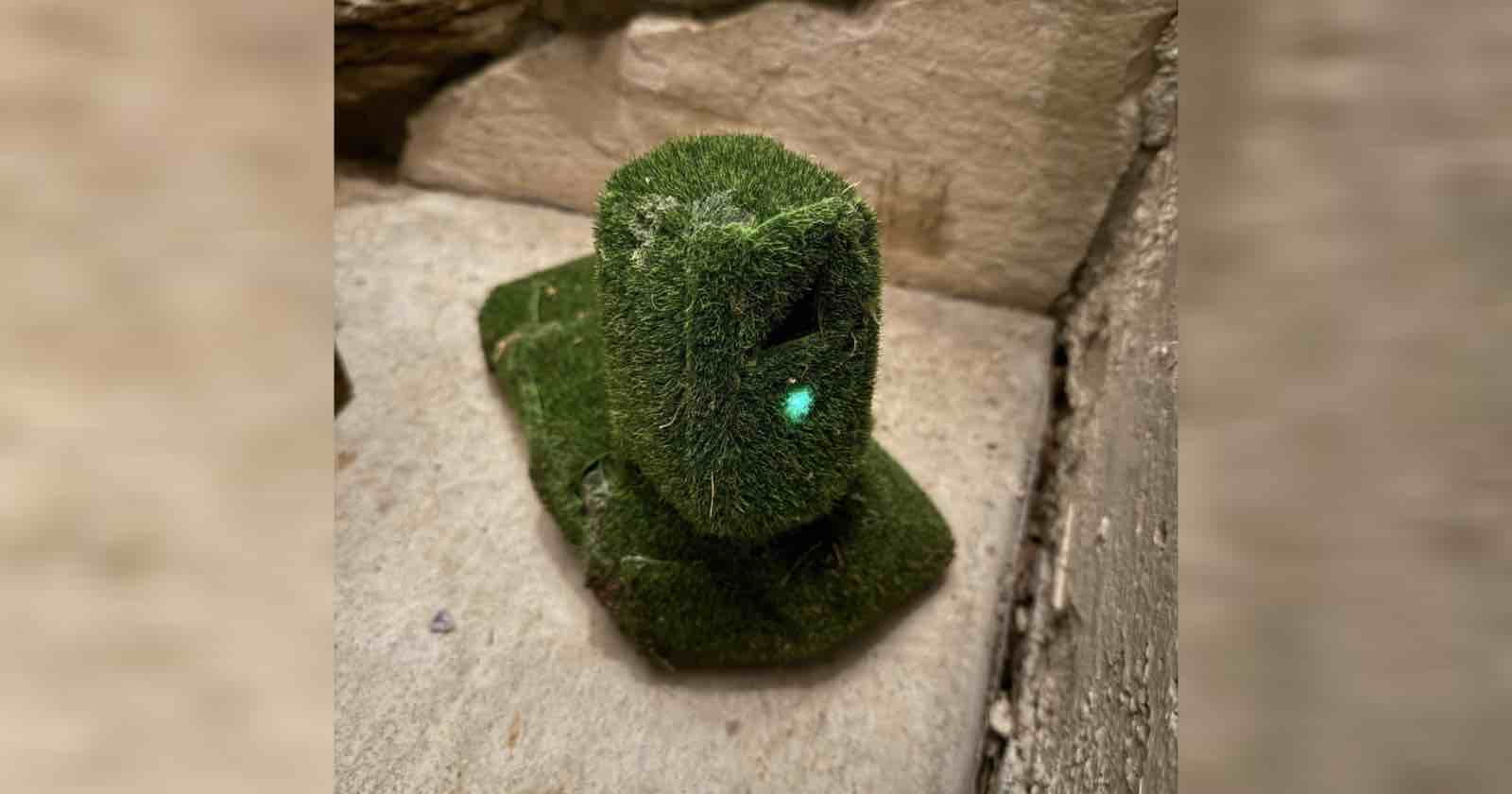 A small sculpture resembling an Easter Island Moai, covered in artificial grass, stands on a concrete surface against a stone wall. A small, glowing green light is visible on its face.