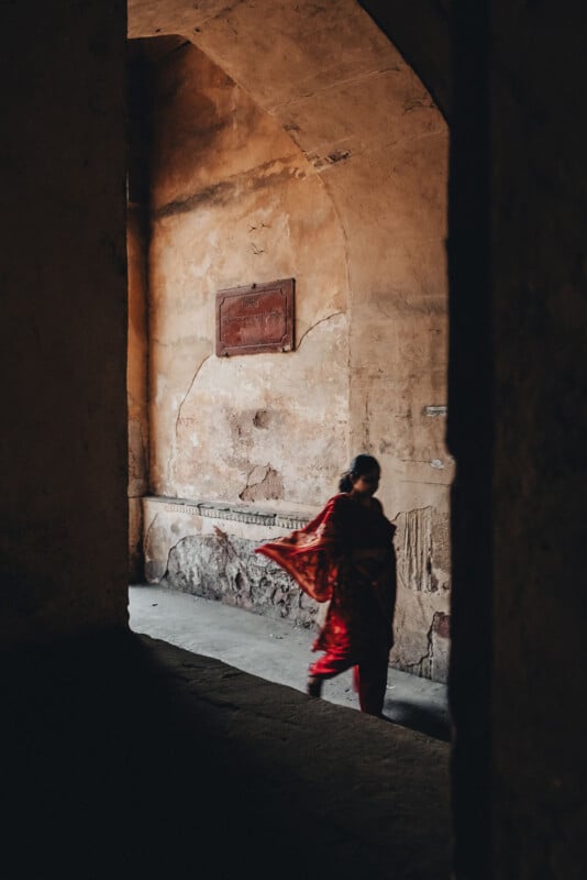 A woman in a red sari walks through a dimly lit, weathered corridor with textured, cracked walls and a high arched ceiling.