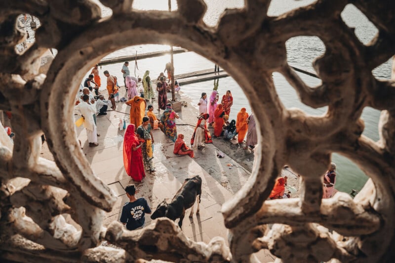 A group of people in colorful clothing gathers at a riverside, seen through an ornate stone lattice. Some stand, others sit or kneel near the water, with a cow in the foreground and bright sunlight illuminating the scene.