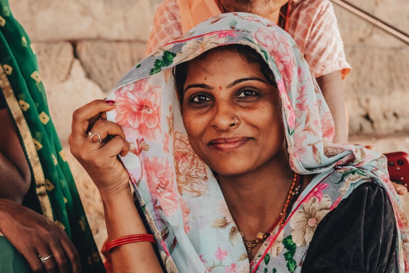 A woman wearing a floral scarf over her head smiles softly at the camera, holding one side of the scarf. She has a nose ring, a red bindi, and colorful jewelry, with a blurred background and another person partially visible.