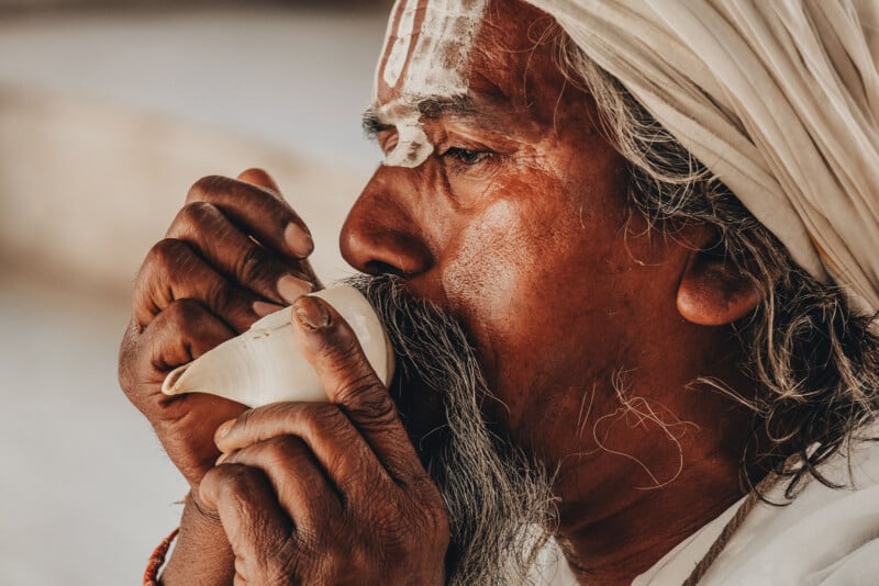 A person with traditional markings on their forehead and wearing a white head covering blows into a conch shell, holding it with both hands, in a close-up shot.