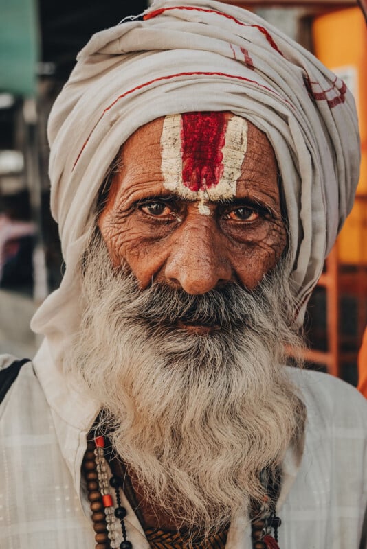 Elderly man with a long white beard and mustache, wearing a white turban and traditional markings on his forehead, dressed in light-colored clothing and beaded necklaces, looking directly at the camera.