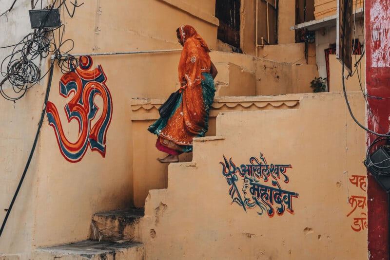 A woman in traditional Indian clothing ascends worn, painted steps beside a yellow wall with colorful Hindi script and an Om symbol, in an urban setting.