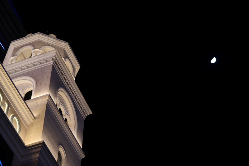 A white, illuminated clock tower against a night sky. The moon is visible in the upper right corner, partially full. The tower features arched windows and a pointed roof.