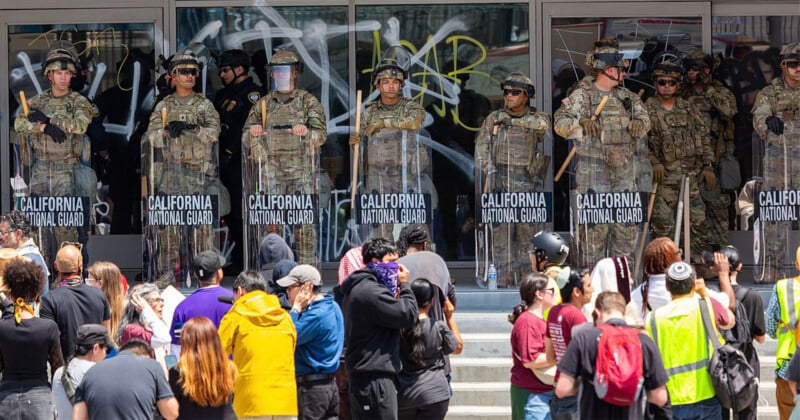 A line of California National Guard troops in riot gear stands in front of a building with glass doors covered in graffiti, facing a crowd of protesters gathered on the steps.