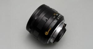 A close-up photo of a black camera lens with gold and white markings, including "50" for the focal length, set against a plain light gray background.