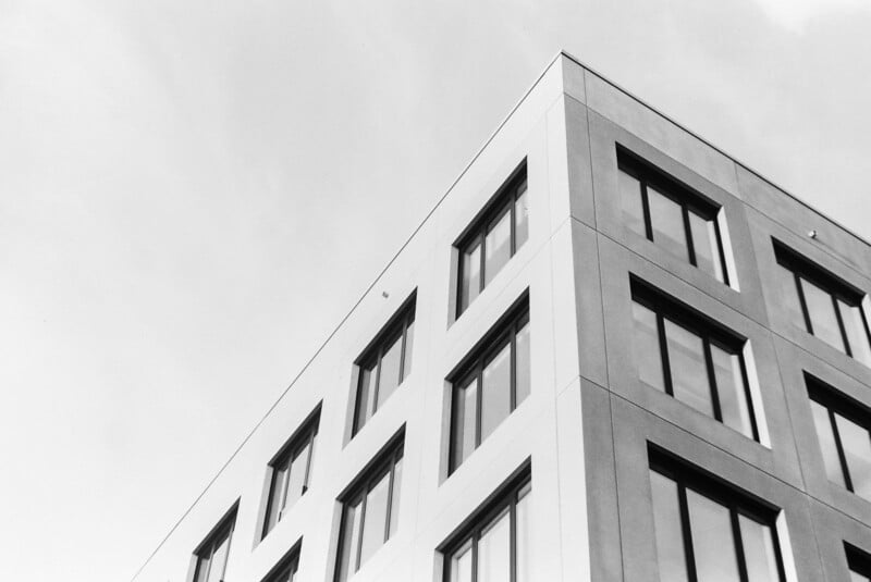 Black and white photo of the corner of a modern, multi-story building with large rectangular windows, viewed from below against a cloudy sky.