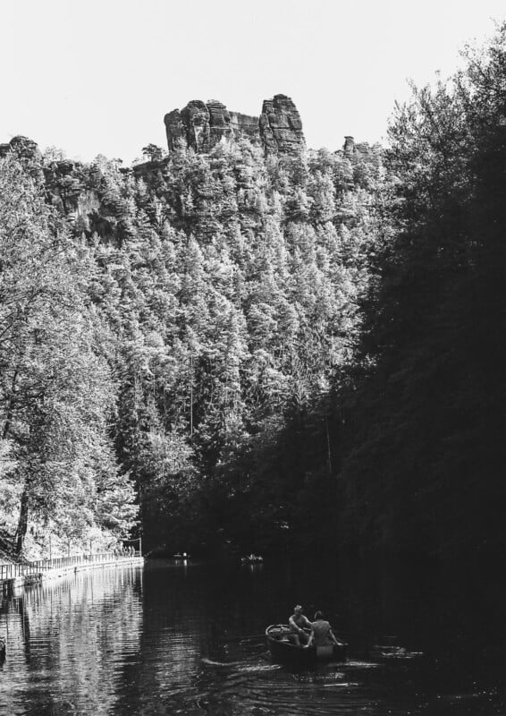 Black and white photo of people rowing a boat on a calm river, surrounded by dense forest, with tall rocky cliffs rising in the background under a bright sky.