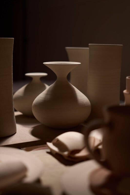A group of unglazed ceramic vases and vessels with smooth, matte surfaces are arranged on a table, illuminated by soft, warm light that creates gentle shadows.