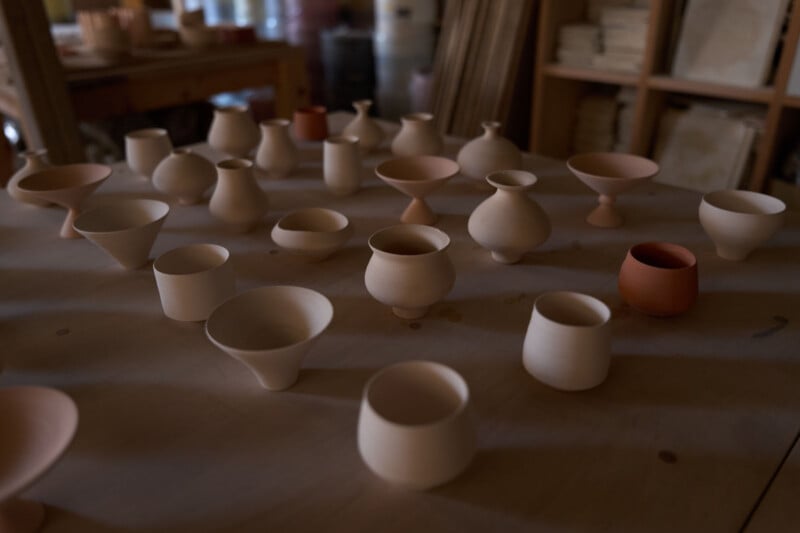 A collection of unglazed ceramic vessels in various shapes and sizes are displayed on a wooden table in a pottery studio, with shelves and tools visible in the background.