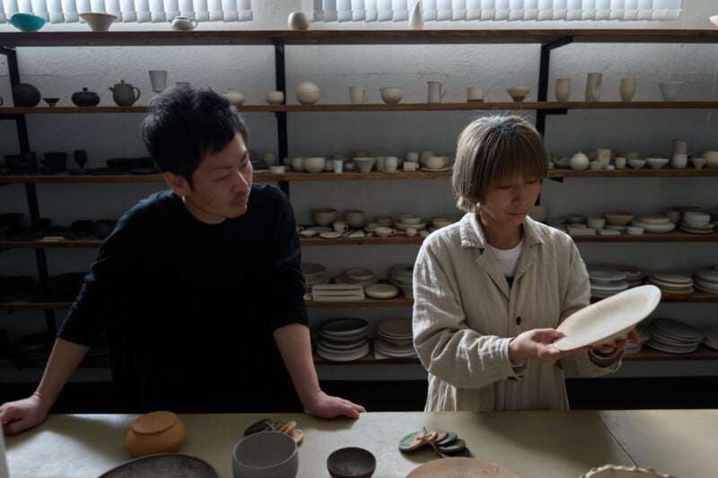 Two people stand in a pottery studio; one examines a ceramic plate while the other looks on. Shelves behind them display various pottery pieces under soft, natural light.
