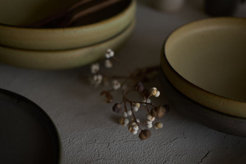 A close-up of ceramic bowls in muted tones, with a small branch of dried white berries resting on a textured, light-colored surface. The lighting is soft and natural, creating a calm, rustic atmosphere.
