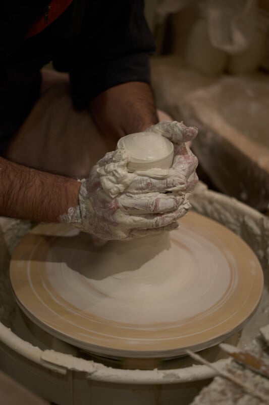 A person with clay-covered hands shapes wet clay on a spinning pottery wheel, focusing on forming a small vessel. The background is blurred, highlighting the pottery-making process.
