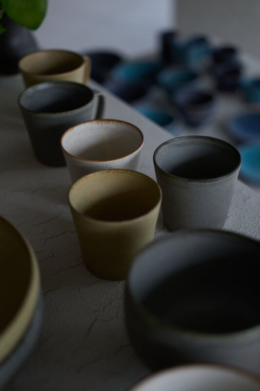 A close-up of several ceramic cups and bowls in muted, earthy tones arranged on a textured light surface, with a blurred background featuring blue and dark-colored objects.