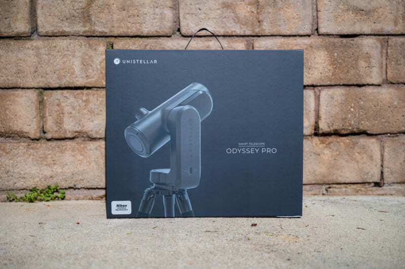 A box for the Unistellar Odyssey Pro smart telescope sits on concrete in front of a stone wall, showing an image of the telescope and its branding on the front.