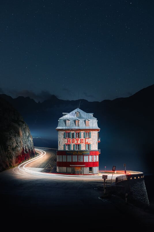 A brightly lit, round hotel stands alone on a mountain road at night, with light trails from passing vehicles curving around it and a starry sky above.