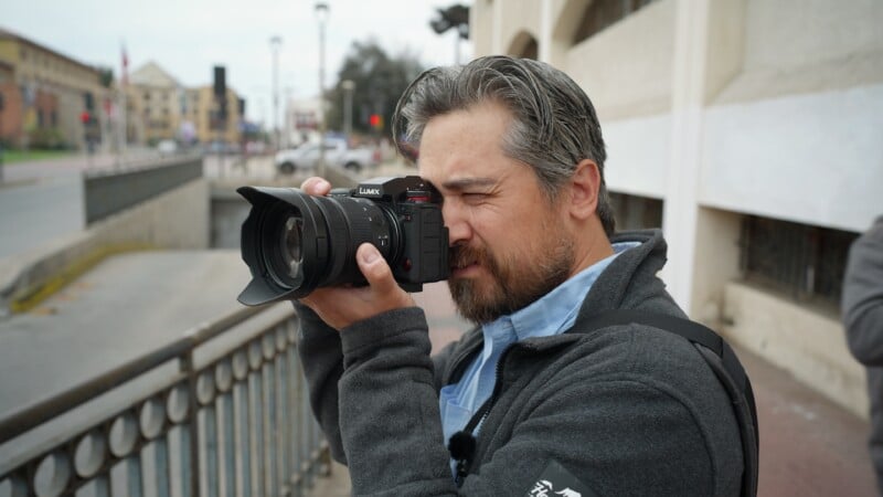 A man with gray hair and a beard is standing outdoors, holding a camera up to his eye as he takes a photo. He is wearing a dark jacket and appears focused, with a blurred city street in the background.