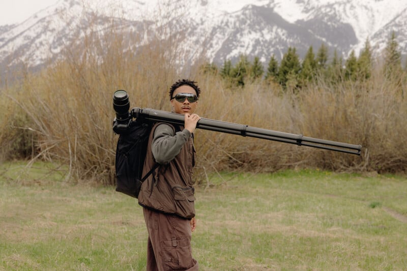 A person wearing sunglasses carries a large camera tripod and bag on their shoulder, standing outdoors in a grassy field with snow-capped mountains and trees in the background.
