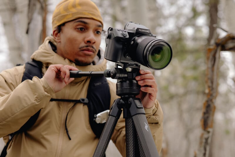 A person in a tan jacket and yellow hat adjusts a camera on a tripod in a forest setting. The scene is outdoors, with blurred trees in the background.