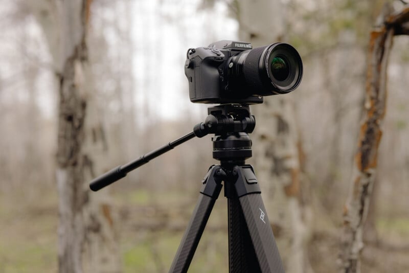 A black Fujifilm camera with a zoom lens is mounted on a tripod outdoors in a forested area, with blurred trees in the background and soft, natural lighting.