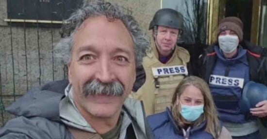 A close-up of four people, three wearing "PRESS" vests and helmets. Two are standing and one is kneeling. The person in front has curly hair and a mustache, smiling at the camera. Two others are wearing masks, and one holds a helmet.
