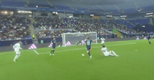 A soccer player in a navy blue uniform prepares to shoot the ball toward the goal as a defender in white slides on the grass. The stadium is filled with spectators watching the action.