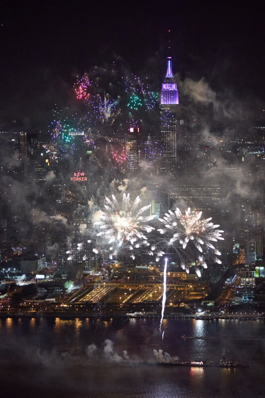 Colorful fireworks explode over the New York City skyline at night, with the illuminated Empire State Building in the background and smoke drifting above the Hudson River in the foreground.