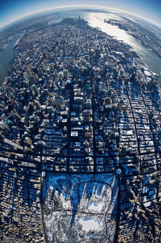 Aerial view of Manhattan, New York City in winter, with snow covering Central Park and rooftops. Sunlight reflects off the rivers and tall buildings, creating a dramatic cityscape under a clear blue sky.