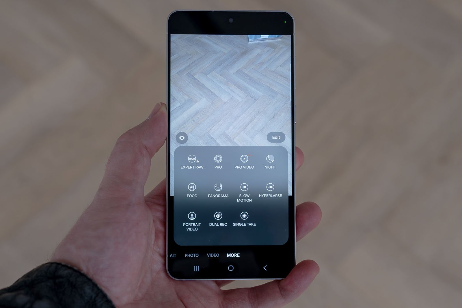A hand holds a smartphone displaying its camera app interface, showing various photography modes like Pro, Night, Panorama, Slow Motion, and Hyperlapse. The background is a light wood floor.