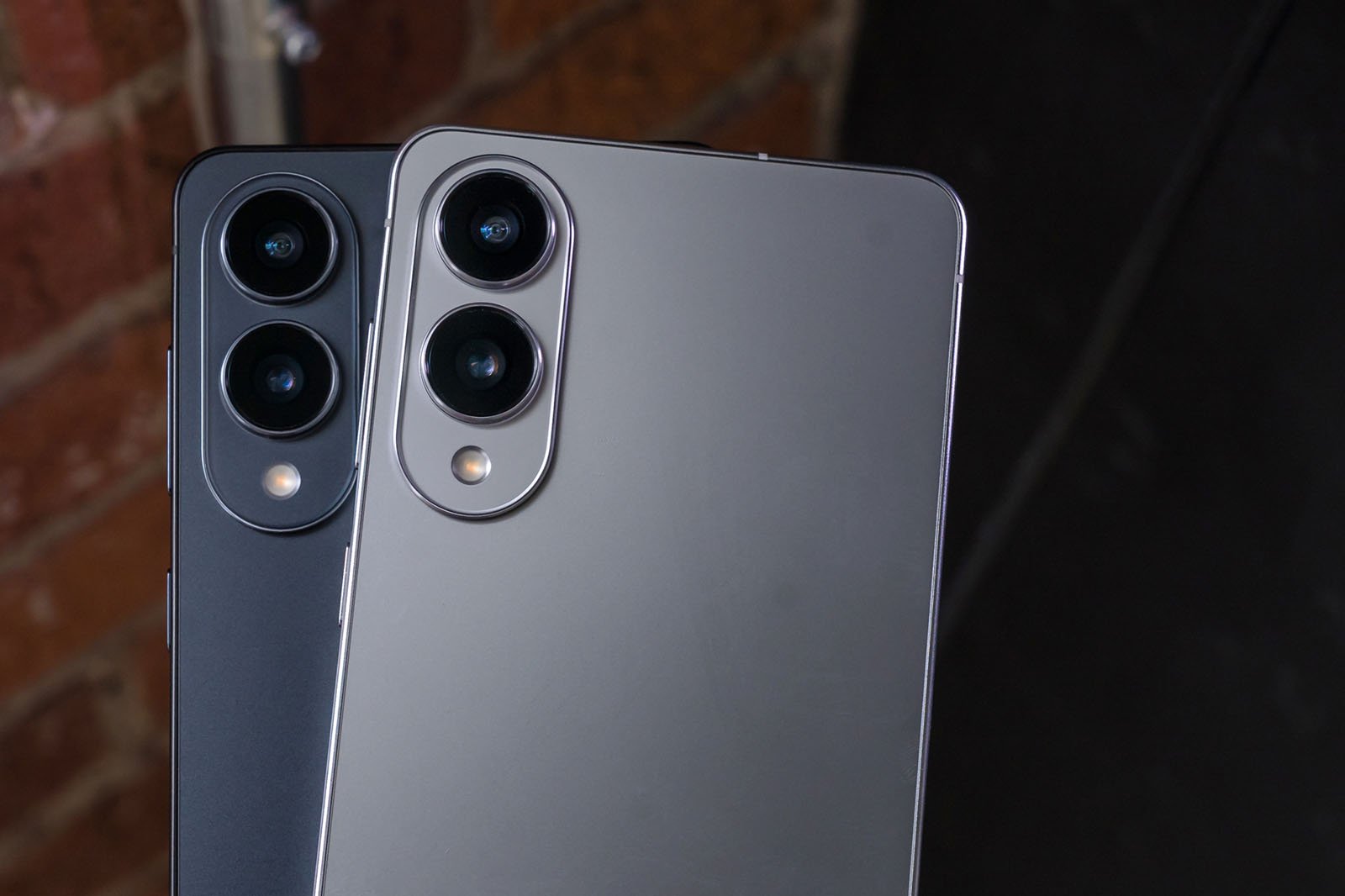 Two modern smartphones, one black and one silver, are shown overlapping with their backs facing up. Each phone has two rear cameras aligned vertically in the top left corner. A brick wall is blurred in the background.