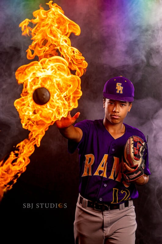 A young baseball player in a purple RAMS uniform throws a ball surrounded by flames, creating a dramatic fire effect. He wears a purple cap and glove, with a smoky, colorful background behind him.