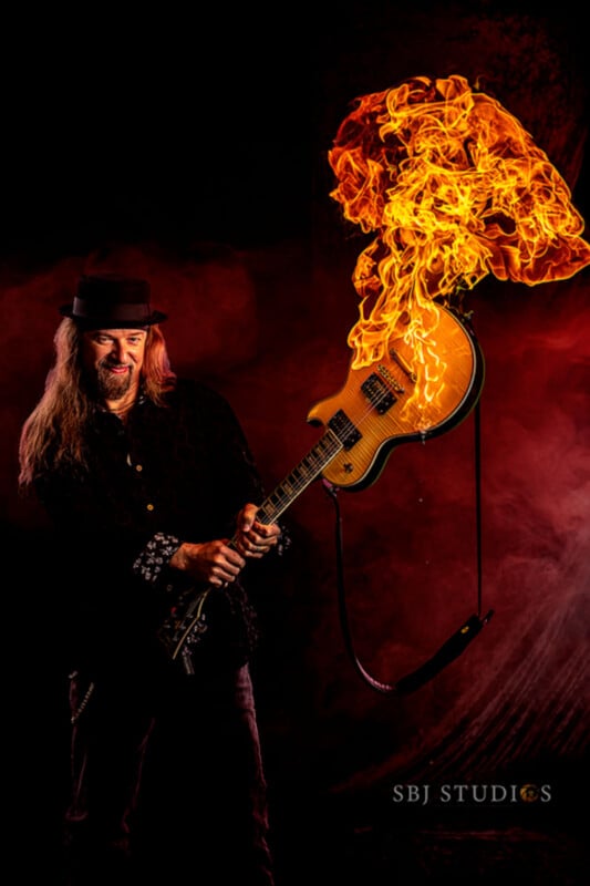 A man with long hair and a hat plays an electric guitar that has flames shooting out from it, set against a dramatic dark red and black background. "SBJ STUDIOS" is written in the bottom right corner.