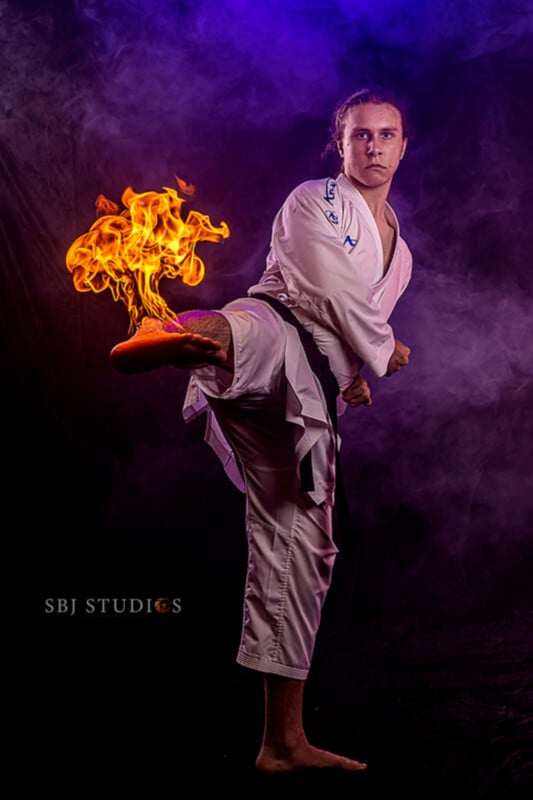 A martial artist in a white uniform and black belt performs a high kick, with flames digitally added to his raised foot. Purple smoke and dramatic lighting enhance the dynamic scene.