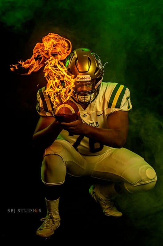 A football player in a gold helmet and white uniform kneels, holding a football with flames rising from it, surrounded by dramatic green and yellow smoke.