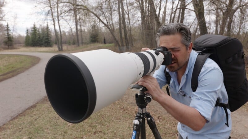 A person with a backpack uses a large telephoto camera lens on a tripod to take photos outdoors in a wooded park area on a cloudy day.