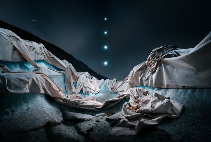 Sheets of white fabric drape over icy terrain at night, illuminated by soft blue light. Five bright stars are aligned vertically in the dark sky above the dramatic, surreal landscape.