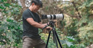 A person stands outdoors among green foliage, adjusting a large camera with a telephoto lens mounted on a tripod, preparing to take a photograph.