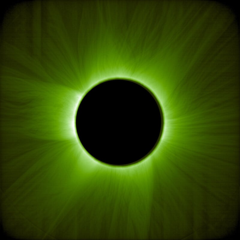A total solar eclipse shown in green tones, with the dark silhouette of the moon blocking the sun, and bright rays of the solar corona radiating outward against a black background.